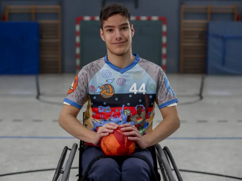 Ein junger Mann im Rollstuhl umfasst einen Ball auf seinem Schoß und schaut freundlich in die Kamera. Im Hintergrund ist ein Handballtor aufgebaut.