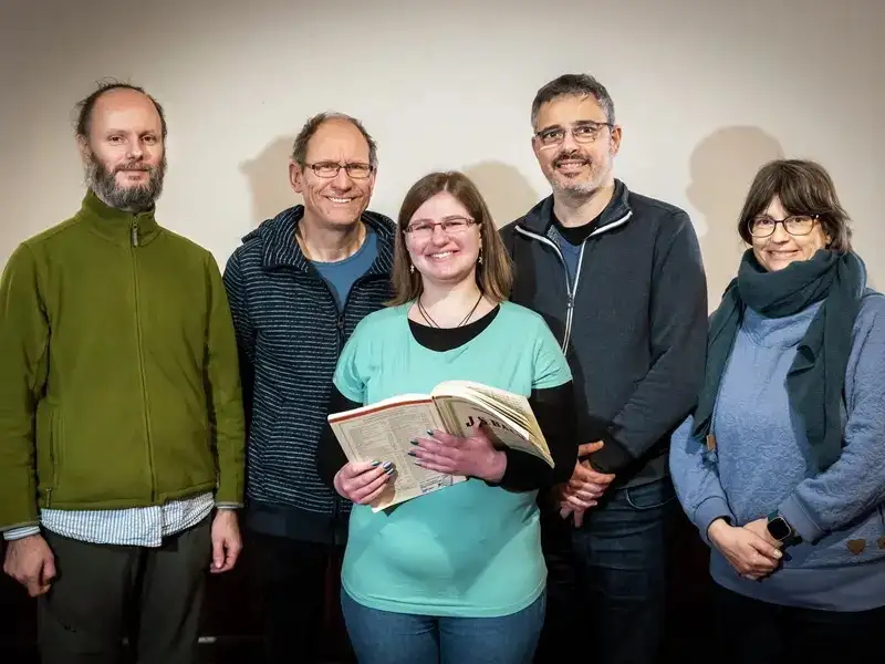 Fünf Personen lächeln in die Kamara. Eine von ihnen hat ein aufgeschlagenes Notenbuch in der Hand.