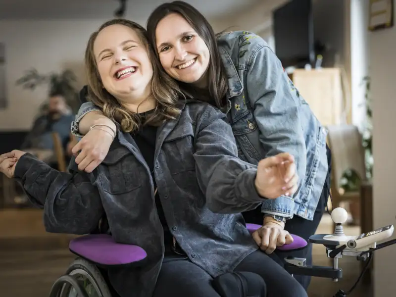 Zwei junge Frauen lachend Arm in Arm, eine von beiden im Rollstuhl