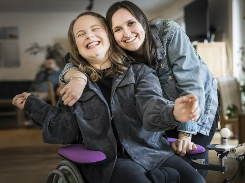 Zwei junge Frauen lachend Arm in Arm, eine von beiden im Rollstuhl