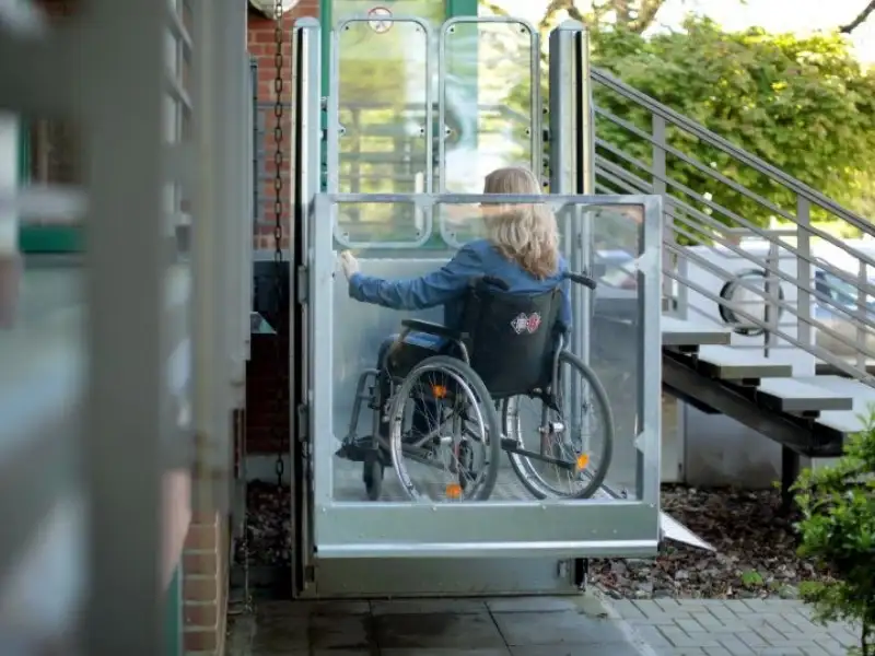 Eine Frau sitzt im Rollstuhl in einem gläsernen Außenaufzug, daneben führt eine Treppe ins Gebäude.