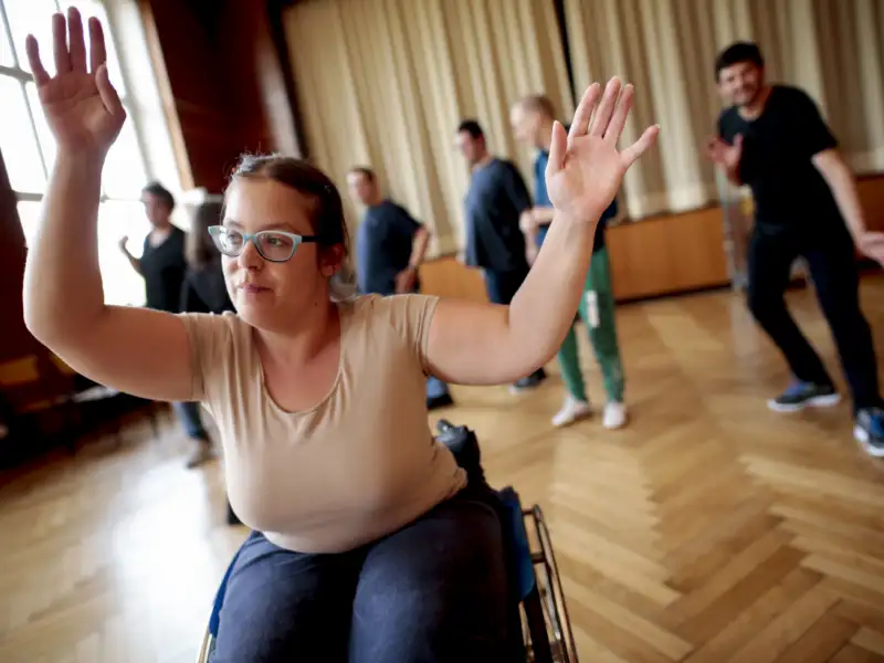 Eine junge Frau im Rollstuhl in einem Übungssaal. Sie hebt beide Arme hoch. Hinter sind weitere Tanzende zu sehen.