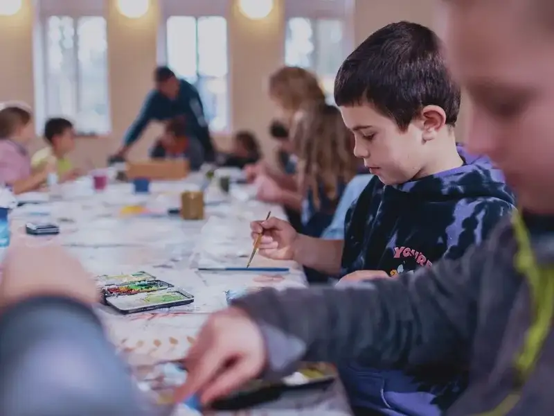 Ein kleiner Junge sitzt mit vielen anderen Kindern an einem langen Tisch und malt.