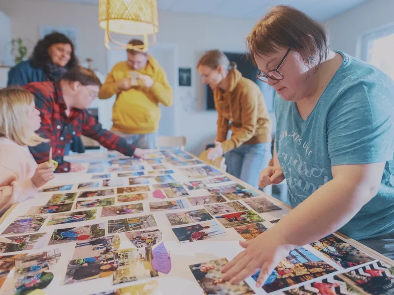 Fünf Personen mit und ohne Behinderung stehen rund um einen Tisch, auf dem viele Fotos liegen
