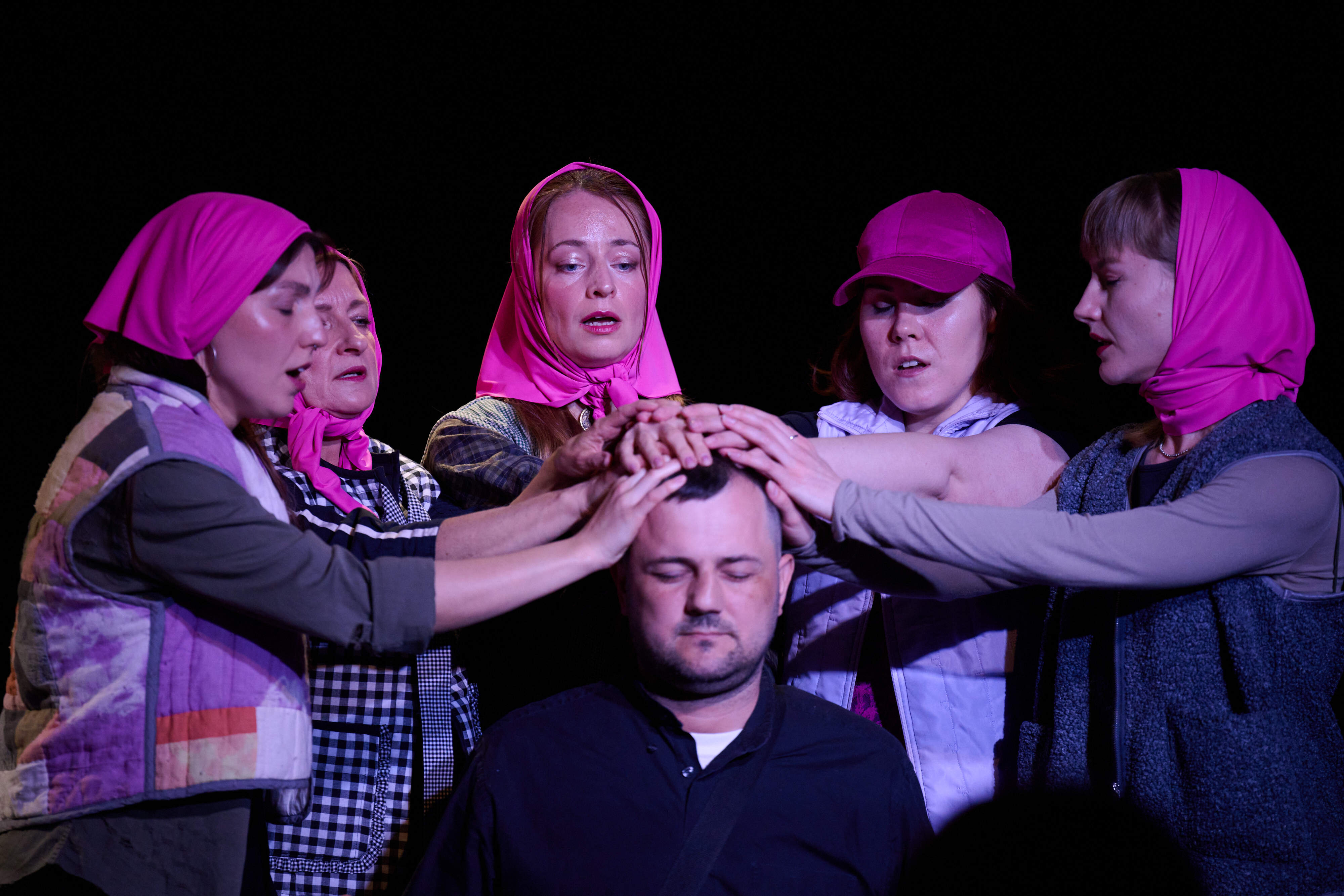 Theaterszene: 5 Frauen mit pinkem Kopftuch legen die Hand auf den Kopf eines Mannes