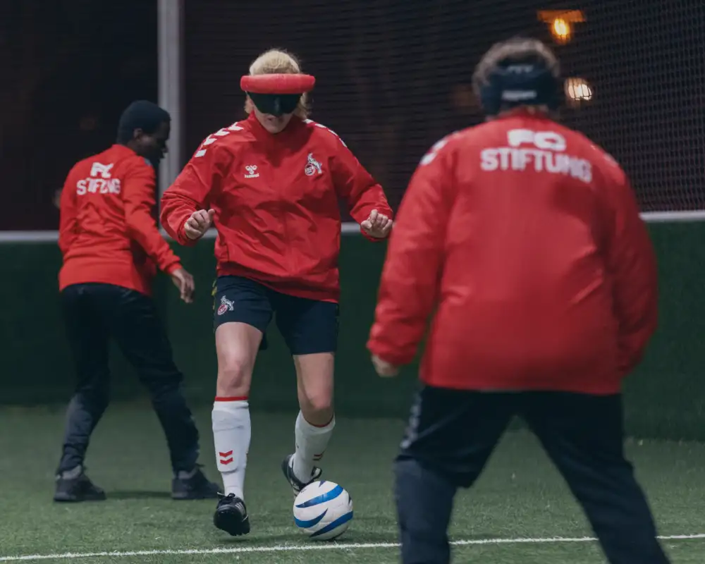 Aktion Mensch - 20.01.2026 - Aufbau eines Blindenfußballteams des 1. FC Köln und der FC Stiftung auf der Sportanlage des Unisports in Köln mit Spieler*innen von 11 bis 47 Jahren. Spielerin Suse Stedtfeld gehört zu den Leistungsträgerinnen des Teams. Für sie ist Fußball mehr als ein Hobby. Sie spielt im Deutschen Blindenfußballteam der Frauen und trainiert jede Woche bei der FC-Stiftung in Köln mit anderen blinden und sehbehinderten Spielerinnen und Spielern. Die 30-Jährige zeigt uns, wie der Fußball für blinde Menschen funktioniert. Blindenfußballspielerin Suse Stedtfeld (Mitte) mit Dunkelbrille beim Dribbling während eines Trainingsspiels mit ihren Mitspieler*innen im Blindenfußballteam der FC Stiftung Köln auf der Sportanlage des Unisports in Köln.