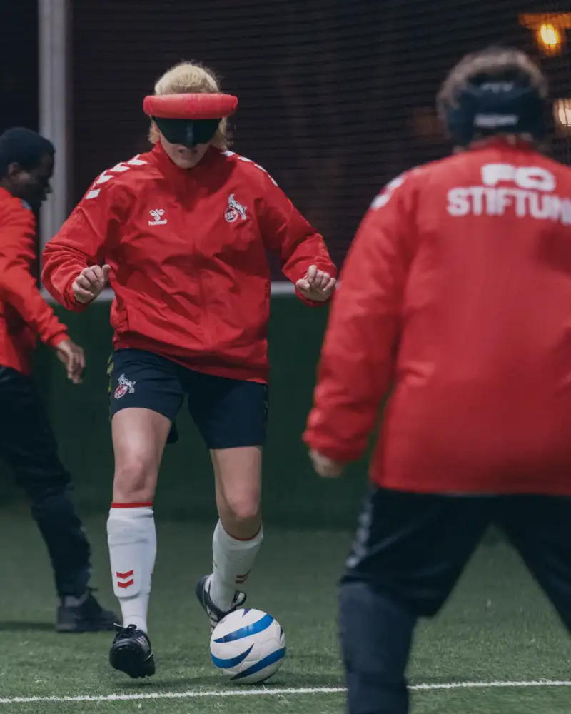 Aktion Mensch - 20.01.2026 - Aufbau eines Blindenfußballteams des 1. FC Köln und der FC Stiftung auf der Sportanlage des Unisports in Köln mit Spieler*innen von 11 bis 47 Jahren. Spielerin Suse Stedtfeld gehört zu den Leistungsträgerinnen des Teams. Für sie ist Fußball mehr als ein Hobby. Sie spielt im Deutschen Blindenfußballteam der Frauen und trainiert jede Woche bei der FC-Stiftung in Köln mit anderen blinden und sehbehinderten Spielerinnen und Spielern. Die 30-Jährige zeigt uns, wie der Fußball für blinde Menschen funktioniert. Blindenfußballspielerin Suse Stedtfeld (Mitte) mit Dunkelbrille beim Dribbling während eines Trainingsspiels mit ihren Mitspieler*innen im Blindenfußballteam der FC Stiftung Köln auf der Sportanlage des Unisports in Köln.