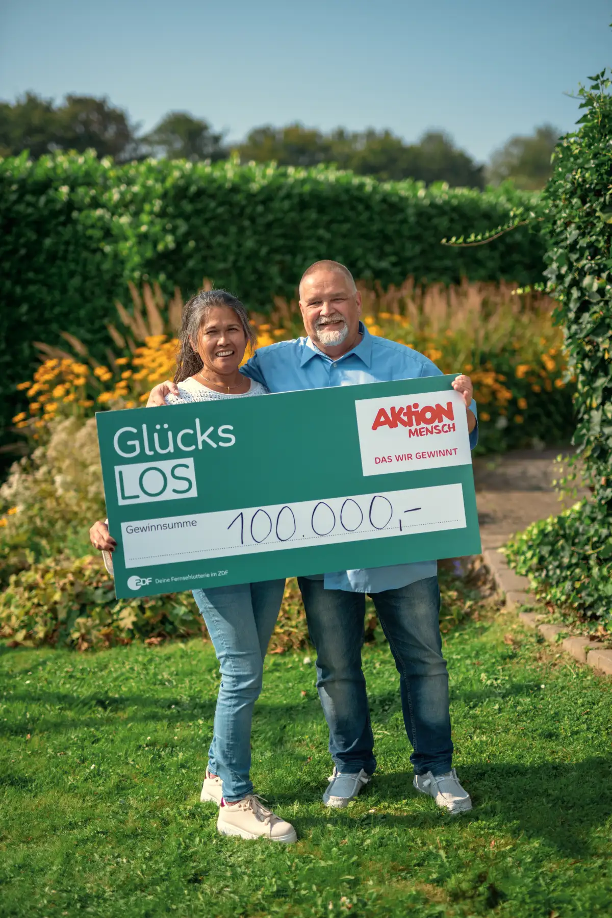 Ralf und seine Partnerin haben 100000 € bei der Aktion Mensch gewonnen und halten freudestrahlend das Glückslos.