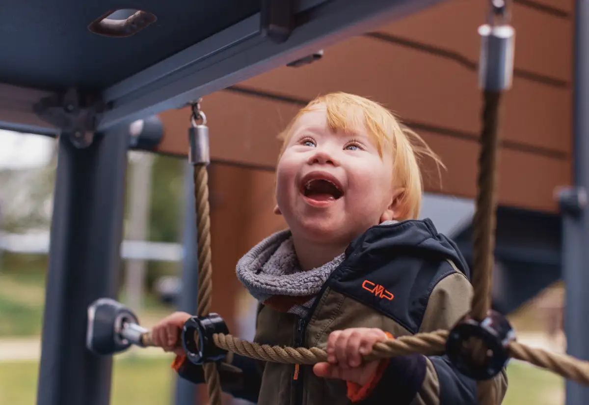 Ein kleines Kind mit Down-Syndrom strahlt vor Freude beim Klettern auf dem integrativen Spielplatz Stück zum Glück Timmel