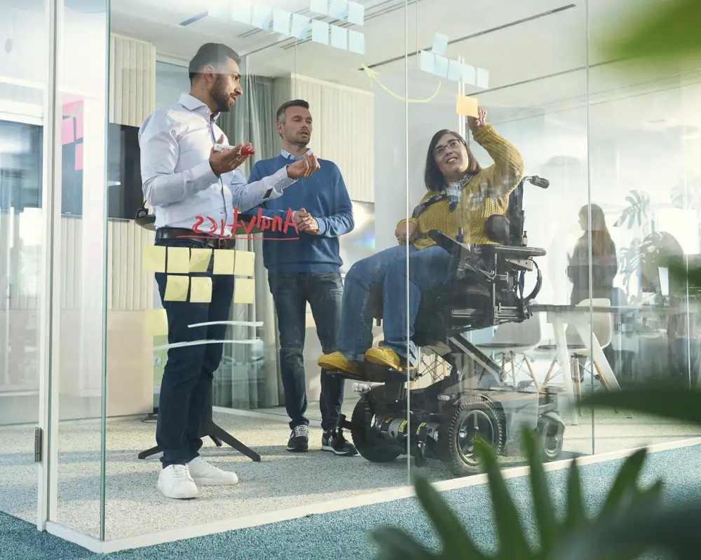 Drei Personen arbeiten in einem Besprechungsraum, eine Frau sitzt im Rollstuhl.