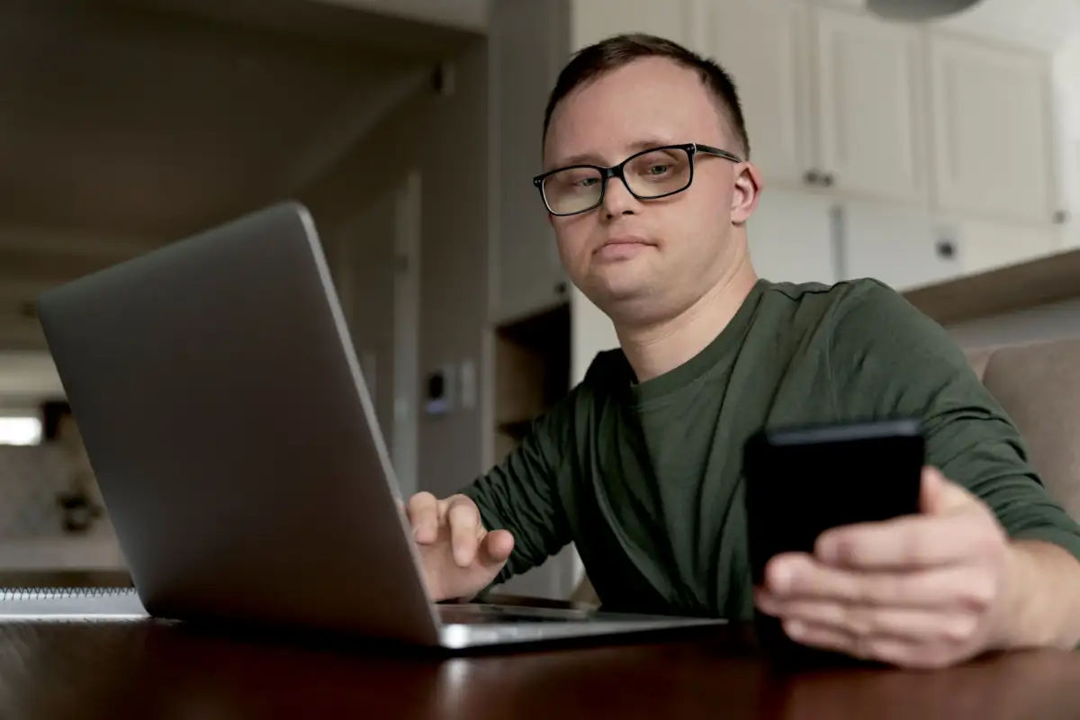 Ein junger Mann mit Downsyndrom sitzt vor einem Laptop. Gleichzeitig hat er ein Handy in der Hand.