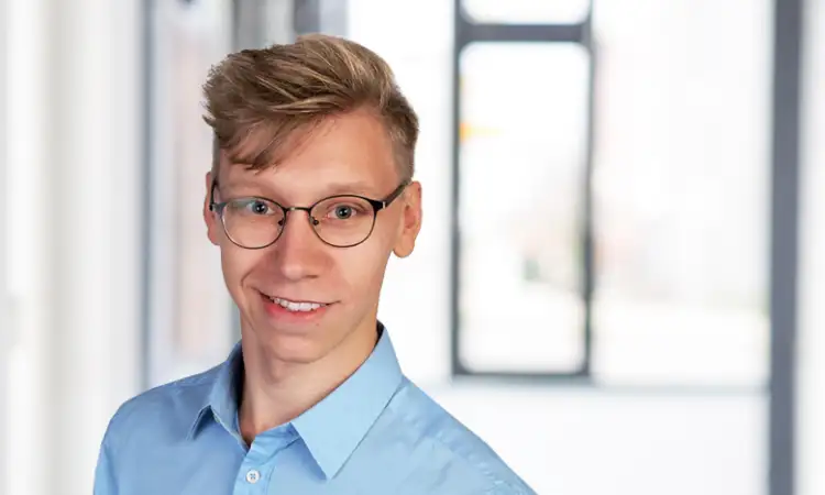 Philipp Oettel, ein junger Mann mir blonden Haaren und Brille trägt ein blaues Hemd und lächelt in die Kamera.