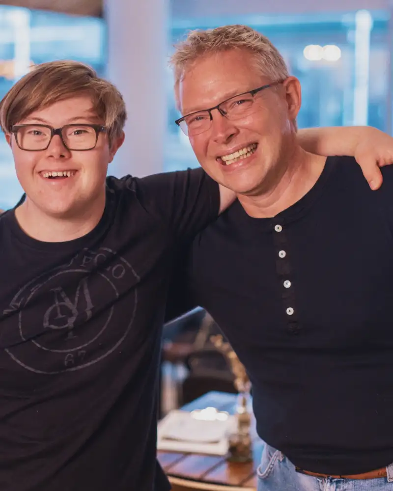 Ein Mann mit Down-Syndrom und ein Mann mit Brille haben einen Arm um die Schultern des anderen gelegt.