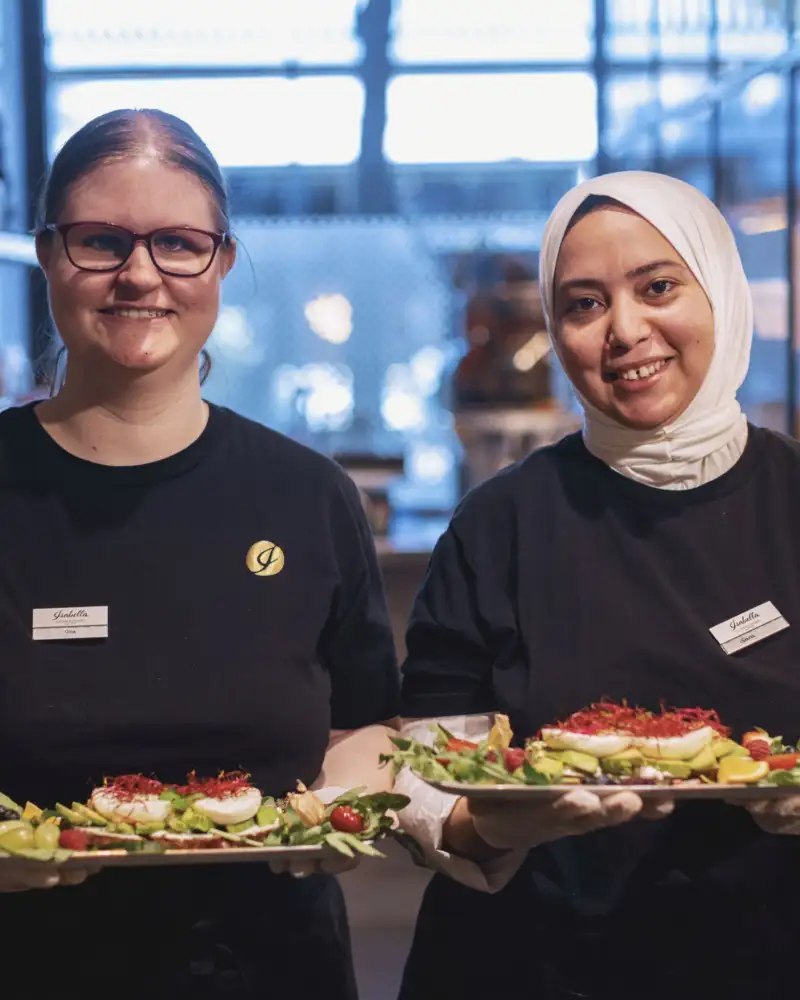 Zwei Arbeitskolleginnen des inklusiven Cafés Isabella in Frankfurt  a. M. aus dem Servicebreich servieren von Gästen bestellte Menüs.