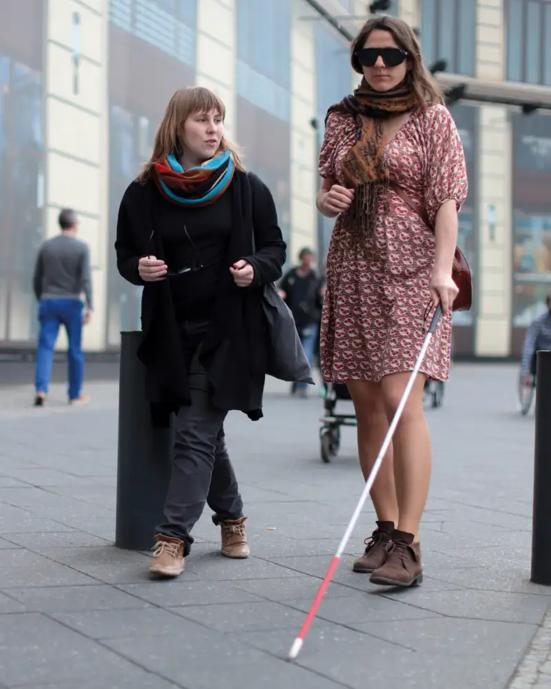 Zwei Frauen gehen durch die Stadt, eine nutzt einen Blindenlangstock.