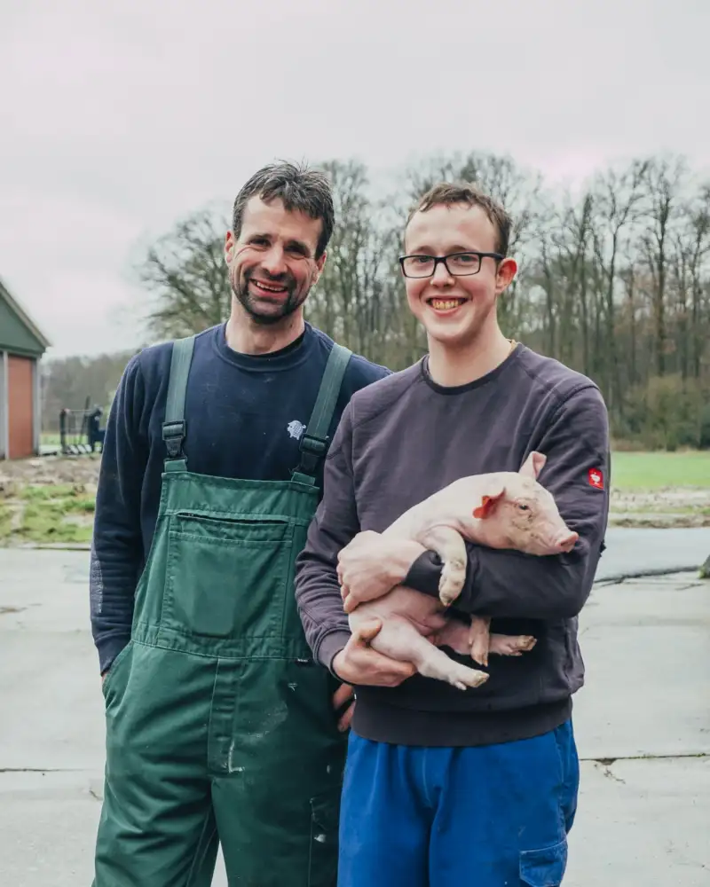 Ein Mann mittleren Alters und ein junger Mann stehen nebeneinander vor einem Bauernhof. Der junge Mann hält ein junges Ferkel auf dem Arm.