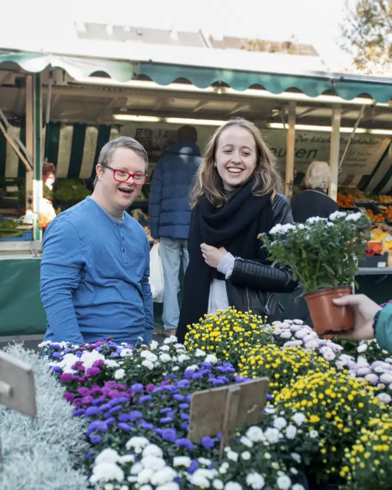 Ein junger Mann mit Downsyndrom und eine junge Frau schauen sich Blumen auf einem Markt an.