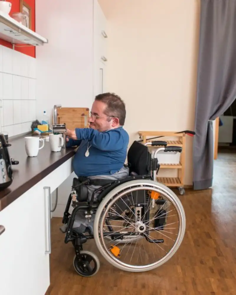 Ein kleinwüchsiger Mann im Rollstuhl bereitet an einer Arbeitsplatte einer Küche einen Kaffee vor.