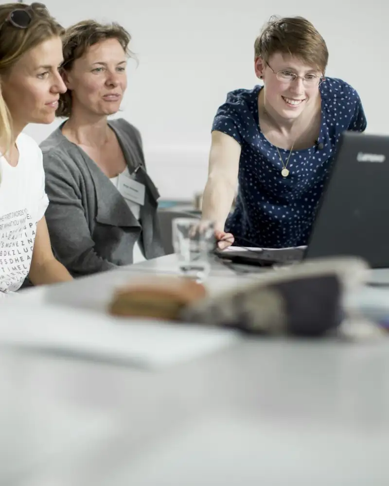 Mehrere Personen sitzen an Schultischen und gucken gemeinsam auf einen Laptop.