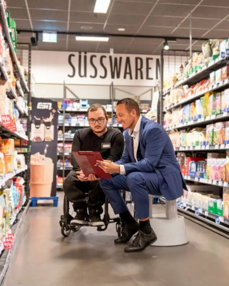 REWE Supermartkbetreiber David Hegemann und sein Auszubildenden Achilleas Tsantekidis, der im Rollstuhl sitzt, vor den warenregalen im Supermarkt.