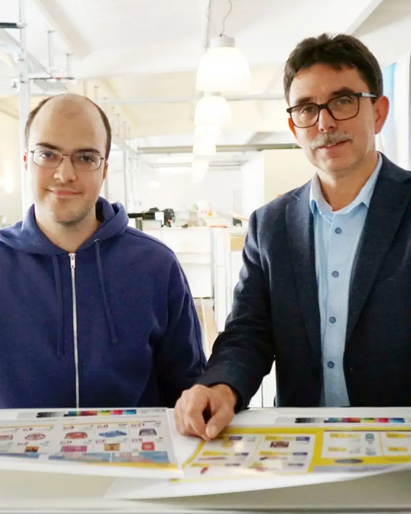 Dennis Niecznik (links) und sein Chef Guido Richter stehen nebeneinander an einem Arbeitstisch der Werbeagentur.