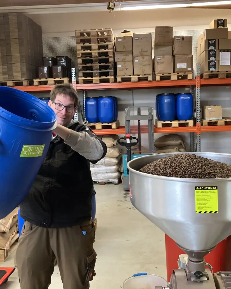 Mitarbeiter Marvin Tutsch schüttet Kaffebohnen aus einem Fass in eine große Kaffeemühle.