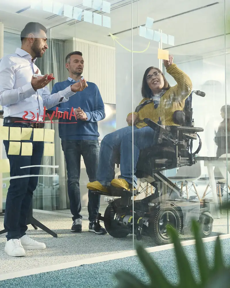 Drei Personen arbeiten in einem Besprechungsraum, eine Frau sitzt im Rollstuhl.