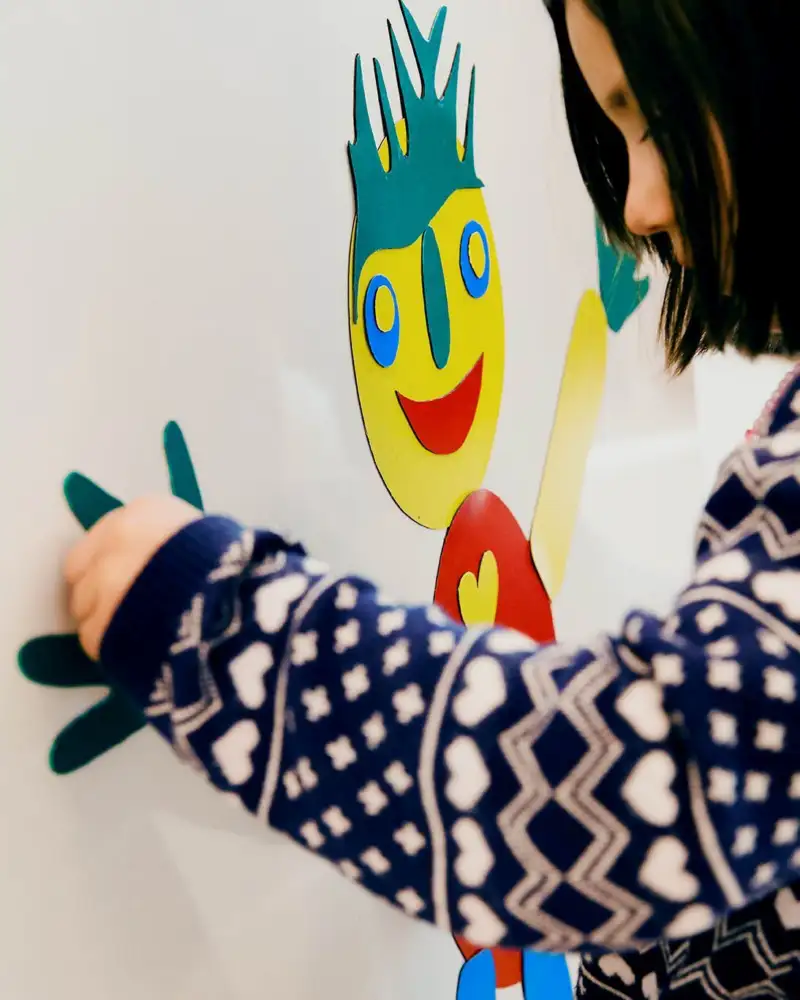 Ein Kind in der Kita klebt eine bunte Figur auf eine Leinwand.