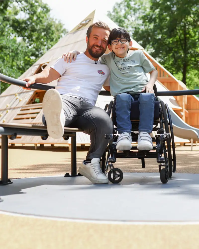 Ein Junge im Rollstuhl und ein kleinwüchsiger Mann sitzen in einem barrierefreiem Spielplatz-Karusell.
