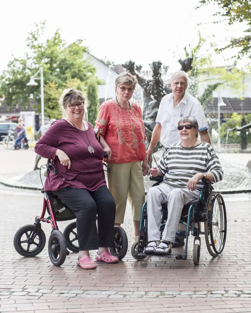 Vier Menschen mit und ohne Behinderung stehen vor einem Brunnen auf einem Platz.