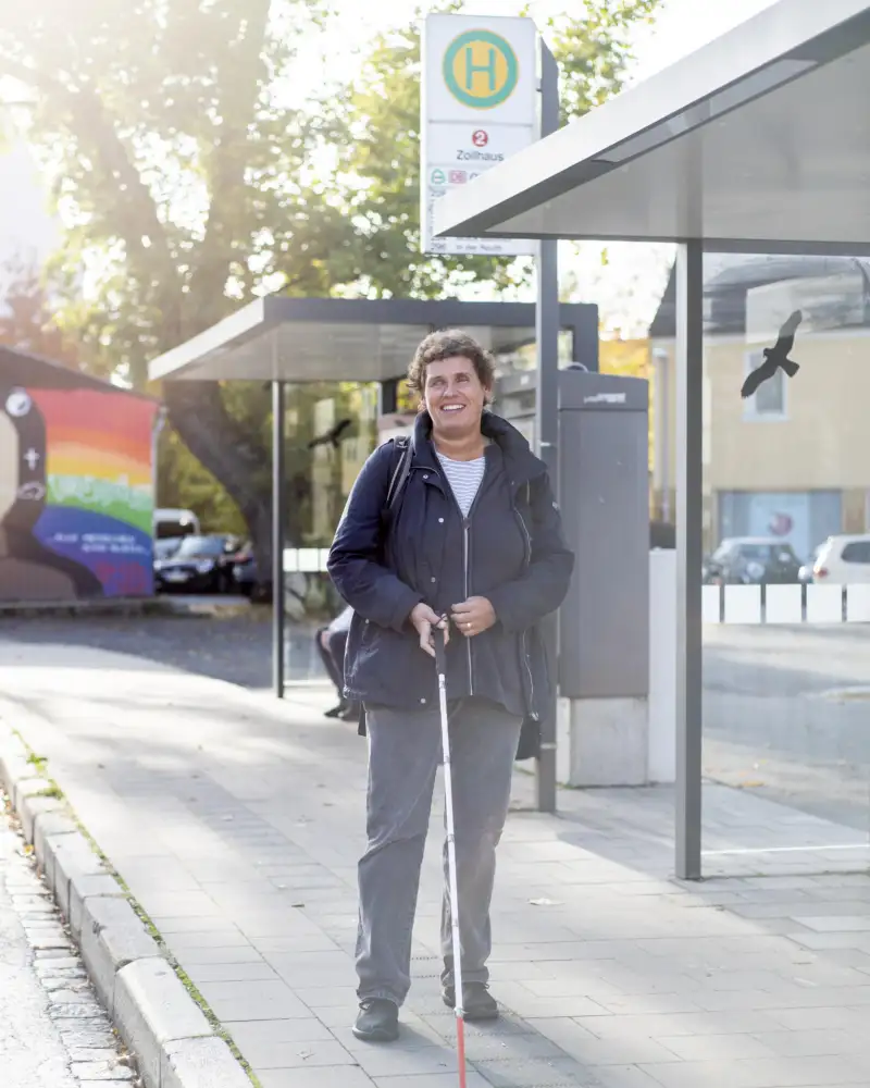 Eine Frau mit Blinden-Langstock steht an einer Bushaltestelle