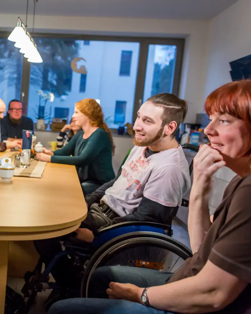 An einem Esstisch sitzen viele verschiedene Menschen mit und ohne Behinderung.