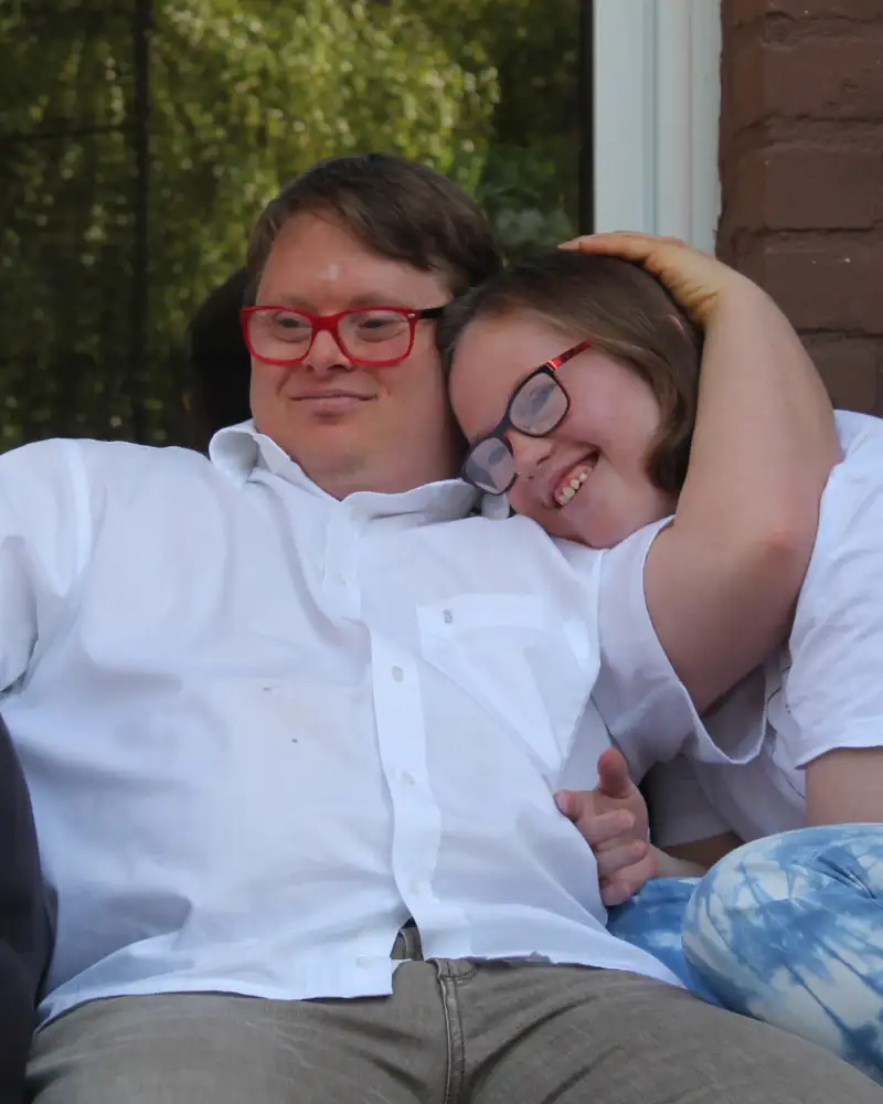 Ein junges Pärchen - beide mit Downsyndrom - sitzen auf dem Sofa. Er hat seinen Arm um sie gelegt und sie kuschelt sich an ihn an.