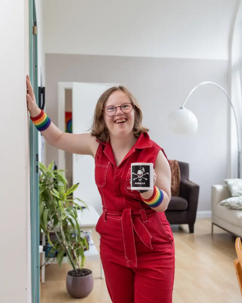 Natalie Dedreux steht in einem roten Jumpsuite in einem Wohnzimmer und lacht in die Kamera. Sie hält sich mit der rechten Hand an der Wand fest.