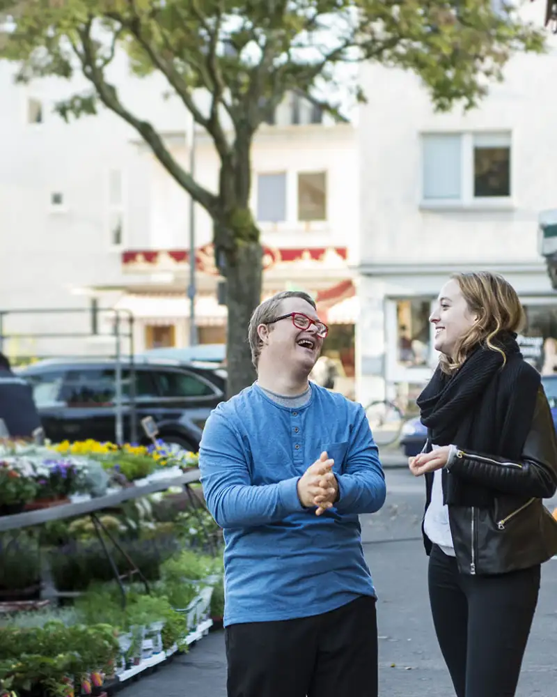 Junger Mann mit Down-Syndrom und eine junge Frau stehen lachend neben einem Blumenstand vom Wochenmarkt