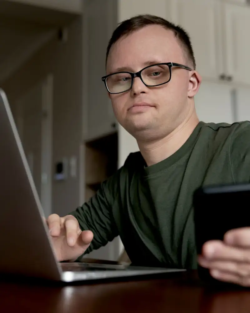 Ein junger Mann mit Downsyndrom sitzt vor einem Laptop. Gleichzeitig hat er ein Handy in der Hand.