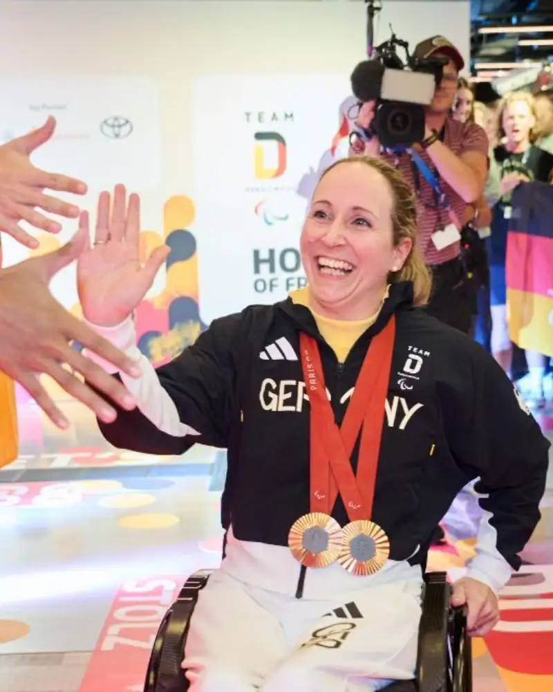 Para Radsportlerin Annika Zeyen-Giles fährt in ihrem Rollstuhl an einer jubelnden Menschmenge im Deutschen Haus Paralympics Paris 2024 entlang. Es werden Deutschlandfahnen geschwenkt. Die Rollstuhlfahrerin klatscht sich mit einem Mann ab.