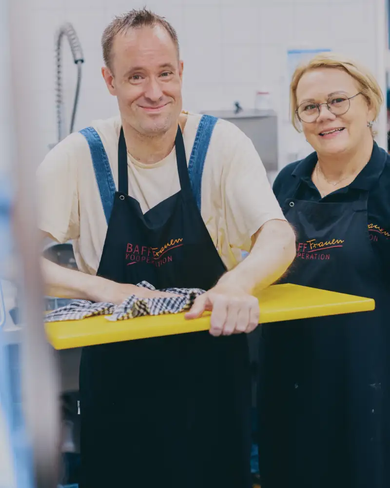 Ein Mann und eine Frau stehen in einer Großküche. Der Mann trocknet ein Schneidebrett ab.