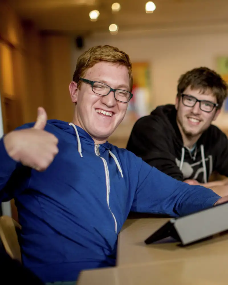 Ein junger Mann mit Behinderung sitzt an einem Tablet und gibt lachend das Daumen-hoch-Zeichen.