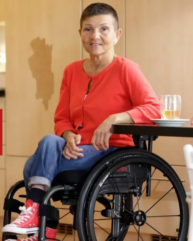 Silvana sitzt im Rollstuhl an einem Tisch. Sie trägt ein rotes Oberteil, eine blaue Jeans und rote Turnschuhe. Neben ihr steht ein Glas mit einem Getränk.