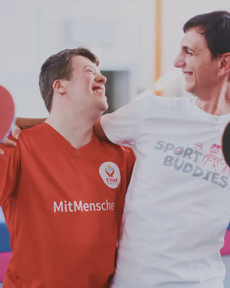 Zwei junge Männer, einer mit Behinderung, einer ohne stehen in einer Sporthalle und halten sich lachend im Arm. Die haben beide einen Tischtennisschläger in der Hand.
