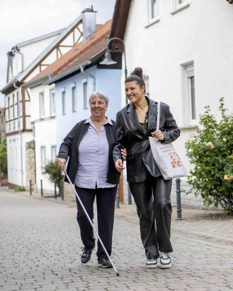 Zwei Frauen laufen eine Straße entlang, eine Frau nutzt einen Blinden-Langstock.