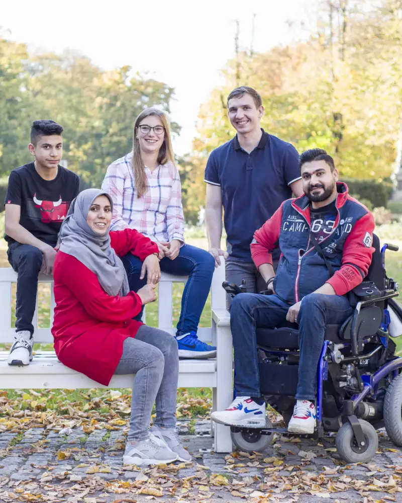 Fünf Menschen sitzen und stehen um eine Bank in einem Garten, ein Mann sitzt im Rollstuhl.