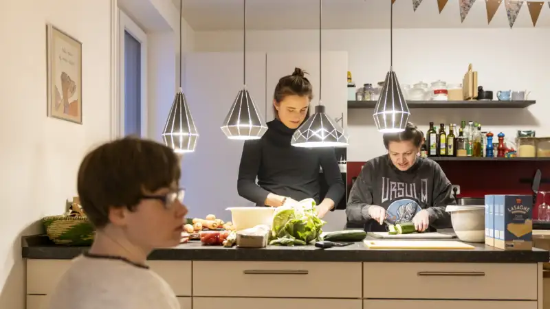 Im Hintergrund schneiden zwei Frauen Gemüse. Im Vordergrund sitzt ein junger Mann.