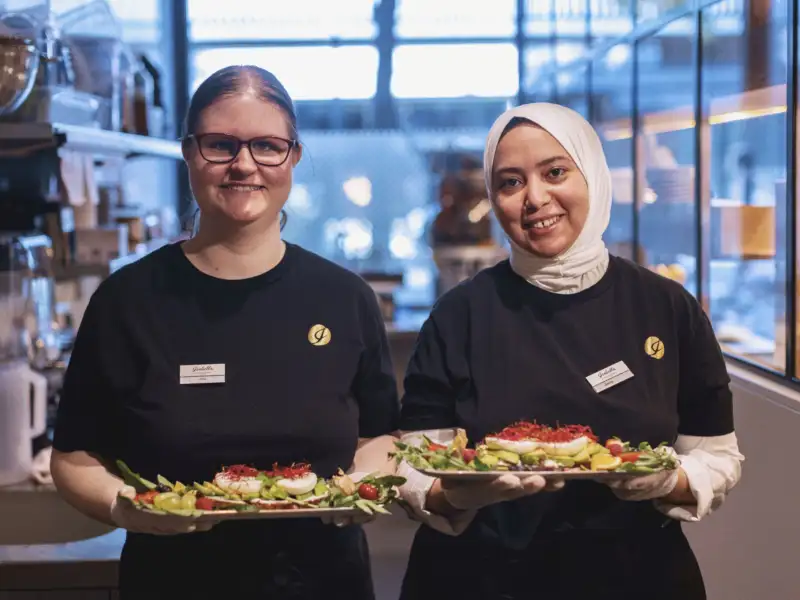 Zwei Arbeitskolleginnen des inklusiven Cafés Isabella in Frankfurt  a. M. aus dem Servicebreich servieren von Gästen bestellte Menüs.