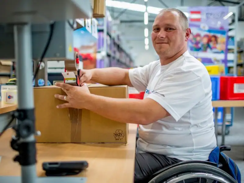 Ein Mann im Rollstuhl sitzt an einem Arbeitstisch und verklebt ein Paket.