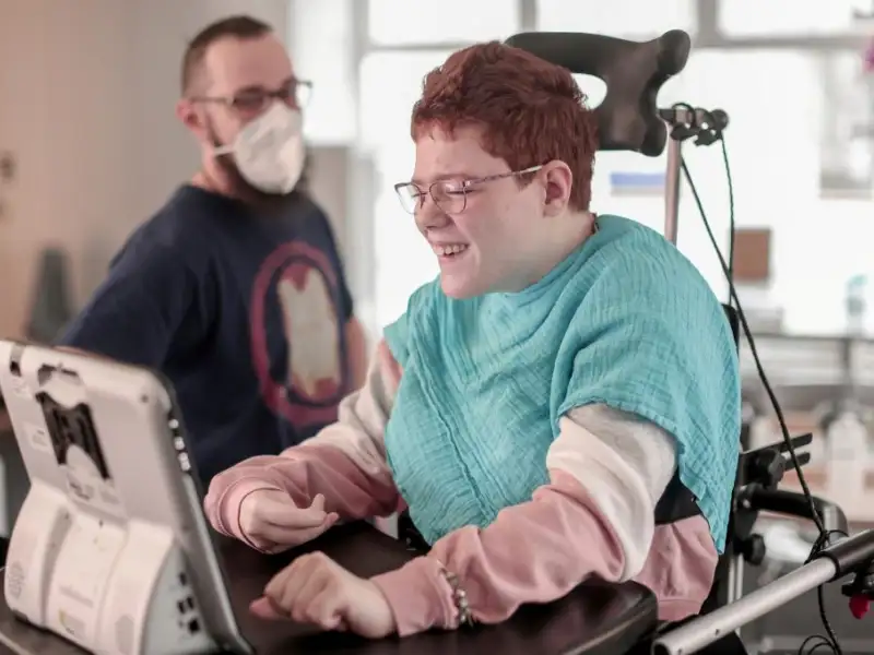 Eine Frau im Rollstuhl arbeitet an einem Sprachkomputer. Im Hintergrund steht ein Mann.