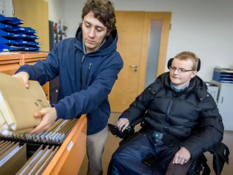 Ein junger mann sitzt im Rollstuhl in einem Büro. Vor ihm steht sein Arbeitsassistent und holt eine Akte aus einem Schrank.