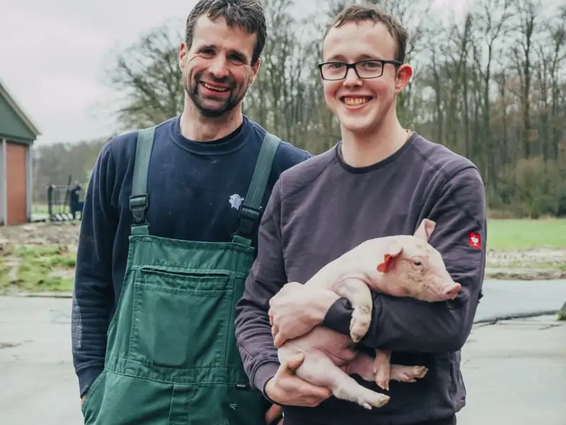 Ein Mann mittleren Alters und ein junger Mann stehen nebeneinander vor einem Bauernhof. Der junge Mann hält ein junges Ferkel auf dem Arm.