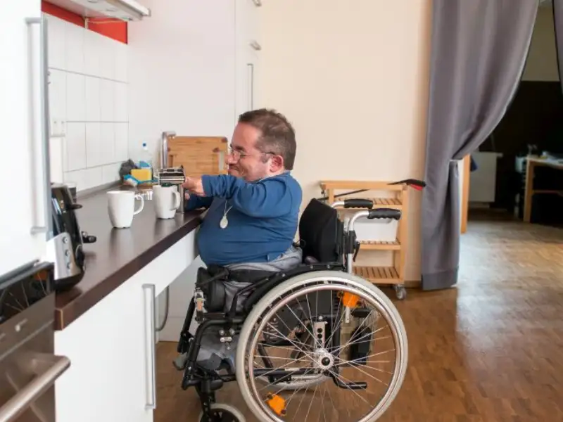 Ein kleinwüchsiger Mann im Rollstuhl bereitet an einer Arbeitsplatte einer Küche einen Kaffee vor.